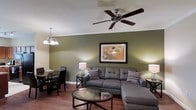 This single-floor space offers a thoughtfully designed interior that blends comfort and functionality. The living area is a standout feature, showcasing sage green accent walls that create a warm, inviting atmosphere. Rich hardwood flooring runs throughout, adding a sense of warmth and continuity to the space. A large sectional sofa anchors the living room, complemented by a glass-top coffee table and tasteful decorative accents. Crown molding lines the ceiling, adding a refined architectural touch, while a ceiling fan with a brushed nickel finish provides both style and practical airflow. Overhead, a classic chandelier adds an elegant lighting element to the adjoining dining area, which features a dark-toned dining set suited for everyday meals or entertaining. Table lamps on either side of the sofa provide layered ambient lighting throughout the space. Framed wall art adds a personalized touch to the décor. The open layout between the living and dining zones creates a seamless flow, making the space feel connected and spacious. Overall, the interior strikes a balance between relaxed living and polished design details.
