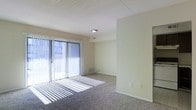 This single-floor space features a open living area finished with wall-to-wall carpeting in a neutral gray-brown tone, providing a comfortable and practical underfoot surface. The room is characterized by its clean, minimalist design, with smooth beige walls and white trim that contribute to a bright, airy atmosphere.

A prominent sliding glass door serves as the focal point of the main living area, fitted with vertical blinds that allow for adjustable light control. When open, the blinds permit abundant natural light to stream in, casting distinctive linear shadows across the carpeted floor. The door also provides direct access to an outdoor area, offering views of the surrounding exterior.

The ceiling features a textured tile finish and is equipped with a centrally located flush-mount light fixture, complemented by a visible air vent for climate control. A baseboard heating unit is positioned beneath the sliding door, ensuring efficient temperature regulation near the primary glass surface.

The space transitions through a partial wall divider, suggesting a defined layout between functional zones. Electrical outlets are conveniently positioned along the walls. Overall, the space presents a straightforward, functional design well-suited for comfortable everyday living.