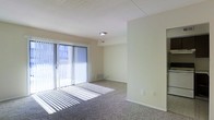 This single-floor space features a open living area finished with wall-to-wall carpeting in a neutral gray-brown tone, providing a comfortable and practical underfoot surface. The room is characterized by its clean, minimalist design, with smooth beige walls and white trim that contribute to a bright, airy atmosphere.

A prominent sliding glass door serves as the focal point of the main living area, fitted with vertical blinds that allow for adjustable light control. When open, the blinds permit abundant natural light to stream in, casting distinctive linear shadows across the carpeted floor. The door also provides direct access to an outdoor area, offering views of the surrounding exterior.

The ceiling features a textured tile finish and is equipped with a centrally located flush-mount light fixture, complemented by a visible air vent for climate control. A baseboard heating unit is positioned beneath the sliding door, ensuring efficient temperature regulation near the primary glass surface.

The space transitions through a partial wall divider, suggesting a defined layout between functional zones. Electrical outlets are conveniently positioned along the walls. Overall, the space presents a straightforward, functional design well-suited for comfortable everyday living.