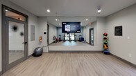This well-appointed fitness studio is designed to support a wide range of workout routines in a clean, contemporary setting. The space features light wood-style flooring throughout, complemented by neutral gray and blue accent walls that create a focused, energizing atmosphere. Recessed lighting and a ceiling fan ensure the room remains bright and comfortable during exercise sessions.

At the heart of the studio is a row of Matrix-brand spinning bikes, ideal for high-intensity cycling workouts. A prominent floor-to-ceiling wall mirror spans one side of the room and is equipped with a mounted display screen, enabling guided fitness classes and virtual training sessions. Additional equipment includes a rack of colorful kettlebells in varying weights, stability balls, a BOSU balance trainer, and medicine balls on a dedicated rack, offering versatile training options.

Natural light filters in through large windows and glass-paneled doors on multiple sides of the room, providing views of the surrounding outdoor landscape. A glass door connects the studio to an adjacent interior space, while exterior doors offer convenient direct outdoor access. The overall layout is open and uncluttered, maximizing usable floor space for stretching, bodyweight exercises, or group fitness activities. This studio is thoughtfully equipped to accommodate a variety of fitness levels and training styles.