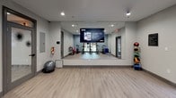 This well-appointed fitness studio is designed to support a wide range of workout routines in a clean, contemporary setting. The space features light wood-style flooring throughout, complemented by neutral gray and blue accent walls that create a focused, energizing atmosphere. Recessed lighting and a ceiling fan ensure the room remains bright and comfortable during exercise sessions.

At the heart of the studio is a row of Matrix-brand spinning bikes, ideal for high-intensity cycling workouts. A prominent floor-to-ceiling wall mirror spans one side of the room and is equipped with a mounted display screen, enabling guided fitness classes and virtual training sessions. Additional equipment includes a rack of colorful kettlebells in varying weights, stability balls, a BOSU balance trainer, and medicine balls on a dedicated rack, offering versatile training options.

Natural light filters in through large windows and glass-paneled doors on multiple sides of the room, providing views of the surrounding outdoor landscape. A glass door connects the studio to an adjacent interior space, while exterior doors offer convenient direct outdoor access. The overall layout is open and uncluttered, maximizing usable floor space for stretching, bodyweight exercises, or group fitness activities. This studio is thoughtfully equipped to accommodate a variety of fitness levels and training styles.