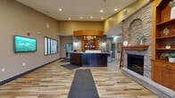 Located at 502 Boulder Dr in Duluth, Minnesota, this expansive single-story community amenity center offers a comprehensive range of facilities across a generously sized floor plan. The building's exterior features stone and brick accents, complemented by well-maintained green lawns, a fenced outdoor sport court, and ample parking.

At the heart of the interior is a welcoming reception area distinguished by a substantial dark wood front desk with marble-toned countertops, warm wood shelving, and a striking stacked-stone accent wall — setting an elevated tone throughout. Natural light filters in through wood-trimmed windows across many rooms.

The fitness center is a standout feature, boasting a warm tongue-and-groove wood ceiling, ceiling fans, rows of treadmills and elliptical machines, free weights, and a mirrored stone accent wall. Adjacent to it, an indoor pool and spa area is enclosed beneath a vaulted wood ceiling, providing a serene aquatic retreat.

A comfortable lounge area offers plush sectional seating, hardwood flooring, and a large projection screen mounted on a stacked-stone wall — ideal for gatherings. A casual dining room with wood-framed windows and light hardwood floors provides additional communal space.

Support areas include well-appointed locker rooms with tile walls and wood benches, and a wide corridor featuring built-in wood cubbies and seating — practical and functional for a high-traffic community facility.