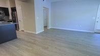 This single-floor space offers a bright, clean, and inviting atmosphere defined by its thoughtful design and neutral aesthetic. The open main living area is finished with light hardwood-style flooring that runs throughout, lending a warm yet modern feel to the space. Soft, neutral gray walls provide a versatile backdrop that complements a wide range of furnishing styles and décor choices.

The layout flows naturally, with a doorway visible from the main area leading to an adjacent room, suggesting a well-connected and functional floor plan. White baseboards provide a crisp contrast to both the flooring and walls, adding a polished finishing touch throughout. Ceiling-mounted HVAC vents indicate a central air system, contributing to year-round comfort.

The space is bathed in even, ambient lighting, creating a welcoming and airy environment. The uncluttered, move-in-ready condition of the interior highlights the quality of the finishes and the generous sense of openness within the layout. Overall, this space presents a practical yet stylish environment well-suited for a variety of uses.