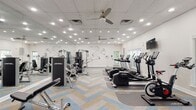 Located at 1623 Parkridge Cir in Crofton, Maryland, this single-floor fitness center is a well-appointed community gym designed to support a comprehensive workout experience. The space is generously sized, offering ample room for both cardio and strength training activities without feeling crowded.

The cardio section features a row of Life Fitness treadmills and elliptical machines, each equipped with individual display consoles and positioned alongside large windows that invite natural light and views of the surrounding greenery. Multiple wall-mounted flat-screen televisions are positioned throughout for entertainment during workouts.

The strength training area is outfitted with a full selection of Life Fitness selectorized machines, a cable crossover station, adjustable benches, and a well-stocked dumbbell rack spanning a wide range of weights. Large floor-to-ceiling mirrors line one wall, enhancing the sense of space and allowing users to monitor their form.

A standout design element is the bold athletic mural painted directly on the white walls, adding an energetic, motivational aesthetic to the space. The flooring features a striking herringbone-patterned carpet in neutral tones with blue accents, lending a modern, polished look. Ceiling fans and recessed lighting ensure the space remains well-lit and comfortable. French glass doors connect to an adjacent area, adding architectural charm to the functional environment.
