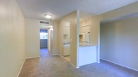 This single-floor residential unit offers a practical and well-organized layout designed for comfortable everyday living. The main living area features wall-to-wall carpeting in a neutral tone, creating a warm and cohesive aesthetic throughout the space. A textured ceiling adds a classic finish overhead, complemented by flush-mount lighting that provides consistent illumination.

The kitchen is positioned adjacent to the living area and is partially enclosed by a half-wall partition, creating a semi-open feel while maintaining a defined cooking zone. White cabinetry lines the upper and lower portions of the kitchen, offering ample storage space, while a white refrigerator and range are included as part of the setup. Light-toned countertops complete the functional kitchen workspace.

A hallway extends from the main living area, lined with the same neutral wall tones and carpeting, leading to additional rooms at the rear of the unit. Natural light filters through a window visible at the end of the hallway, brightening the corridor and hinting at a well-lit room beyond. A centrally located HVAC vent in the ceiling ensures climate control throughout the unit. The overall ambiance is clean, neutral, and move-in ready, suited for those seeking a straightforward and low-maintenance living environment.