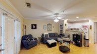 This two-story space offers a welcoming and functional layout, beginning with an open-concept main living area. The living room is generously sized, featuring warm hardwood floors that run throughout and classic crown molding that frames the ceiling, adding a touch of architectural detail. A ceiling fan with integrated lighting provides both comfort and illumination, complementing the overall bright and airy atmosphere of the room.

The living area is well-appointed with ample space for seating arrangements and casual relaxation. A built-in shelving unit along one wall provides convenient display and storage options, adding a practical yet decorative element to the room. The neutral, warm-toned walls create a versatile backdrop suitable for a variety of décor styles.

The living space flows naturally into the kitchen area, which is visible from the main room via a pass-through counter or open partition. The kitchen features white cabinetry and appliances, maintaining a clean and bright aesthetic. The open sightlines between the living and kitchen areas enhance the sense of space and encourage easy interaction between rooms. The second floor adds additional living or sleeping accommodations, making this a practical and well-rounded multi-level home.