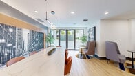 Located at 310 Granello Ave in Coral Gables, Florida, this thoughtfully designed amenity space combines a professional conference area with a relaxed lounge environment on a single floor. The open layout flows seamlessly from one zone to the next, anchored by light hardwood flooring that lends warmth throughout.

At the heart of the space sits a striking elongated marble-top conference table surrounded by ergonomic terracotta-toned chairs, illuminated by a sleek sculptural pendant light. Behind it, a dramatic floor-to-ceiling black-and-white cityscape mural mounted against a warm wood-paneled accent wall adds a bold visual focal point. Recessed ceiling lighting ensures the area is well-lit for productive meetings.

The lounge area features a curated mix of soft gray pod chairs, petite side tables, and vibrant geometric-patterned area rugs that inject color and personality into the space. Lush tropical plants — including tall monstera and leafy potted varieties — are strategically placed throughout, bringing life and a sense of tranquility indoors.

A bold cobalt blue credenza with display surfaces serves as both storage and a design statement. Large glass entry doors invite abundant natural light and provide views of the lush outdoor surroundings. A framed black-and-white photographic artwork completes the sophisticated, gallery-like ambiance of this well-appointed communal space.