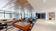 Located at 300 Atlas St in Baltimore's vibrant 21230 corridor, this single-floor lobby and amenity space delivers a refined, contemporary aesthetic that sets a welcoming tone for residents and guests alike. The expansive open-plan layout is anchored by a striking wood-paneled ceiling, its warm tones contrasting elegantly with large-format porcelain tile flooring that stretches across the main circulation areas. Generous floor-to-ceiling windows line the perimeter, flooding the interior with natural light and offering clear views of the surrounding neighborhood streetscape. The lounge area is thoughtfully furnished with a combination of a rich cognac leather sofa, neutral sectionals, and coordinating leather accent chairs, all arranged atop a bold, abstract area rug featuring deep blues, golds, and grays. Rounded stone-clad columns punctuate the space, adding architectural interest while defining distinct seating zones. A communal table with leather-upholstered stools provides a collaborative gathering point. Toward the rear, sleek elevator banks framed by stone wall cladding and white cylindrical columns lead to the upper floors. Glass-partitioned rooms along the side offer additional private or semi-private spaces. The overall ambiance is polished yet inviting, blending modern design sensibilities with warm materials to create a sophisticated and functional common area.