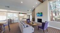 Located at 1700 Via Pacifica in Corona, California, this single-story community clubhouse offers a well-appointed gathering space designed for comfort and functionality. The centerpiece of the main lounge is a dramatic floor-to-ceiling stacked stone fireplace, complemented by a wall-mounted flat-screen television and soaring vaulted ceilings with exposed wood beams. Recessed lighting and a skylight flood the space with brightness, while large windows and glass French doors along the perimeter invite abundant natural light and provide views of the surrounding outdoor area, including palm trees and a pool deck. The lounge is furnished with plush gray sofas and distinctive purple accent chairs arranged around a modern coffee table, creating an inviting social atmosphere. A dedicated dining area features a sturdy wood high-top table with bar-height seating and direct access to the outdoor patio through multiple glass doors. The fully equipped kitchen includes white shaker cabinetry, granite countertops, light hardwood flooring, and stainless steel appliances — including a French door refrigerator, electric range, built-in microwave, and oven. The overall design blends contemporary finishes with warm, residential touches, making this clubhouse a versatile and welcoming amenity space.