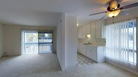 This single-floor residence presents an open-concept layout that seamlessly connects the living area and kitchen. The living space is finished with plush carpeting and benefits from generous natural light streaming through a large sliding glass door fitted with vertical blinds, which opens to an outdoor balcony or patio area with views of surrounding greenery.

A partial dividing wall separates the kitchen from the living space, maintaining an open feel while offering a defined cooking area. The kitchen is outfitted with white cabinetry along the upper and lower sections, providing ample storage. A double-basin sink is positioned beneath under-cabinet lighting, and the space includes a dishwasher. The kitchen floor features a geometric-patterned tile, offering a visual contrast to the carpeted living area.

A ceiling fan is installed in the kitchen area, contributing to air circulation throughout the connected spaces. The overall interior palette is light and neutral, with white walls and bright finishes creating a clean, airy atmosphere throughout the unit.