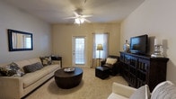 Located at 8439 Dorchester Rd in North Charleston, South Carolina, this single-floor space offers a comfortable and well-appointed layout designed for everyday living. The living area serves as the heart of the space, featuring plush carpet flooring that adds warmth underfoot. A ceiling fan with an integrated light fixture is centrally mounted, providing both airflow and ambient lighting. Natural light filters in through multiple windows dressed with grommet-style curtains, as well as a glass-panel exterior door that brightens the room and offers a connection to the outdoors. The neutral, warm-toned walls create a versatile backdrop that complements a range of furnishing styles. The living area provides ample room for seating arrangements, as evidenced by the space available for a sofa, accent chair with ottoman, and a media console along the wall. The overall ambiance is relaxed and inviting, with a layout that balances openness and functionality. The single-floor design ensures easy accessibility throughout, making this a practical and welcoming space suited for comfortable day-to-day living in the North Charleston area.