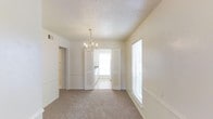 This single-floor space offers a clean, classic aesthetic defined by soft neutral walls, plush carpeted flooring, and bright, airy interiors. The main living area draws the eye upward with a textured popcorn ceiling and a delicate candelabra-style chandelier that serves as a refined focal point, adding a touch of traditional elegance to the room. Chair-rail molding runs along the lower portion of the walls, lending architectural detail and a polished finish throughout. Natural light pours generously into the space through multiple windows fitted with white horizontal blinds, creating a warm and welcoming ambiance. An open passageway connects the living area to an adjacent room, where a glimpse of what appears to be a kitchen is visible — bright and clean, with white cabinetry and a window that further enhances the sense of openness. The layout flows naturally between rooms, offering a cohesive and functional use of space. Louvered bi-fold doors add a classic touch to storage or closet areas, maintaining the consistent traditional style found throughout. Overall, the space presents a bright, well-maintained interior with thoughtful architectural details that contribute to a comfortable and inviting atmosphere.