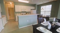 This single-floor space showcases an open-concept layout connecting the kitchen and dining area in a cohesive, functional design. The kitchen is outfitted with white cabinetry that extends to the upper walls, providing ample storage, complemented by white appliances including a range, microwave, and refrigerator. A peninsula with a light-colored countertop serves as a natural divider between the kitchen and dining spaces, offering additional workspace and casual seating potential. The walls throughout are painted in a calming sage green tone, lending the space a relaxed, consistent aesthetic. Flooring transitions from light-toned carpet in the dining area to a harder surface in the kitchen. The dining area comfortably accommodates a dark-finished table and chair set, with a pendant light fixture hanging overhead, adding a warm focal point. A window fitted with blinds allows natural light to filter into the dining space, brightening the area. A secondary dining or breakfast table is also visible near the window, offering flexible seating arrangements. Overall, the space presents a practical and welcoming environment suited for everyday living and dining.