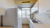 Located at 1445 E Broad St in Columbus, Ohio, this single-floor residential unit offers a practical and functional layout across several well-defined spaces. The entryway greets residents with warm, richly toned parquet hardwood flooring that transitions into the unit's various rooms. The galley-style kitchen is efficiently arranged with white cabinetry, a gas range, a double basin sink, and a full-size refrigerator, all complemented by light wood-look flooring and a window that brings in natural light with views of the surrounding trees. The spacious living area features soft wall-to-wall carpeting, a wall-mounted heating unit, and an opening that connects seamlessly to the rest of the unit. The bedrooms are generously sized, each equipped with ceiling fans, carpeted floors, large windows with outdoor views, and closet storage. Two bathrooms serve the unit — one features classic blue ceramic tile wainscoting, a pedestal sink, mirrored medicine cabinet, and a parquet tile floor, while the second offers cream-toned tile walls with a burgundy accent border, mosaic parquet flooring, a vanity, and a soaking tub with shower. Together, these spaces create a cohesive, livable environment in the heart of Columbus.