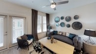 This thoughtfully designed single-floor residence offers a cohesive blend of modern aesthetics and functional living. The open-concept kitchen is a standout feature, showcasing white upper cabinetry paired with wood-toned lower cabinets, a granite-topped island with seating, stainless steel appliances including a French door refrigerator with ice dispenser, a microwave, and a range. Track lighting overhead and a deep undermount sink add both practicality and style, while light hardwood-style flooring ties the space together seamlessly.

The living area is warm and inviting, anchored by a plush charcoal sofa, a light wood coffee table, and a striking gallery wall of decorative metallic disc art. Natural light filters through large windows dressed with elegant floor-to-ceiling charcoal drapes, and a ceiling fan provides year-round comfort.

The bedroom is a serene retreat, featuring plush carpeting, generous windows with coordinating drapes, a ceiling fan, and ample room for a large bed and dresser. The space is accented with contemporary wall art and thoughtful décor details in a calming blue and gray palette.

The bathroom is clean and bright, finished with wood-style flooring, a full tub-shower combination with marble-look tile surround, and crisp white walls, completing this well-rounded and stylish residence.