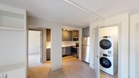This single-floor unit showcases a contemporary design with a neutral color palette, light wood-style flooring, and clean lines throughout. The open kitchen is thoughtfully equipped with gray flat-front cabinetry extending to the ceiling, white countertops, a subway tile backsplash, and a full suite of stainless steel appliances including an electric range, built-in microwave, dishwasher, and refrigerator. A stacked washer and dryer unit is conveniently integrated near the kitchen area, enclosed behind a door for a tidy appearance.

The living area is bright and open, featuring large floor-to-ceiling windows that allow abundant natural light and provide views of the surrounding greenery. Built-in shelving and cabinetry offer practical storage solutions within the main living space. Track lighting runs along the ceiling, adding a modern touch.

The bedroom is well-proportioned, offering a large window with outdoor views, a closet with built-in storage, and light, neutral finishes that contribute to a calm atmosphere. The bathroom features a soaking tub with a white subway tile surround, a floating gray vanity with a white countertop, brushed nickel fixtures, and recessed lighting, completing the unit's cohesive modern aesthetic.