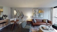 This thoughtfully designed single-floor residence blends contemporary style with mid-century modern sensibilities throughout its well-appointed rooms. The living area serves as the centerpiece of the home, showcasing a tufted brown leather sofa, a patterned area rug, and an oversized abstract painting that adds a bold artistic focal point to the space. Diamond-shaped mirrors adorn one wall, reflecting warm ambient light alongside a coordinating arc floor lamp, creating an inviting and curated atmosphere. The kitchen is efficiently laid out with light maple cabinetry, stainless steel appliances including a refrigerator, range, microwave, and dishwasher, and a generous white countertop breakfast bar that provides both prep space and casual seating. Dark hardwood flooring grounds the kitchen's clean, functional design. The bedroom offers a tranquil retreat, centered around a channel-tufted upholstered headboard flanked by a series of coastal-themed framed prints. A floor-to-ceiling sliding glass door fills the room with natural light and frames striking urban cityscape views. The bathroom is neatly finished with light maple cabinetry, a white tiled tub-shower combination, and a clean vanity with ample storage. Throughout the residence, a consistent cool grey palette and carefully selected furnishings create a cohesive, modern aesthetic.