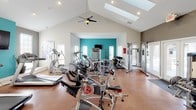 This fitness center is designed to accommodate a wide range of workout needs within a single, open-plan space. The most striking architectural feature is the dramatic vaulted ceiling, which rises to a considerable height and is fitted with skylights that flood the interior with abundant natural light. A ceiling fan assists with air circulation, keeping the environment comfortable during intense workouts. Recessed lighting supplements the natural light, ensuring the space remains well-illuminated throughout the day.

The flooring throughout is a polished, warm-toned surface that is both durable and easy to maintain. The walls are finished in a neutral gray palette, with a bold teal accent wall providing a vibrant focal point. A wall-mounted flat-screen television is positioned on the accent wall for entertainment during cardio sessions.

The equipment selection is extensive and well-organized. The cardio zone includes multiple treadmills, elliptical trainers, stationary spin bikes, and a stair climber. The strength-training area features Precor cable machines, a multi-use weight bench, free weights, and resistance equipment. French doors provide access to the outdoors, further enhancing the airy, open ambiance of this well-appointed fitness facility.