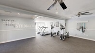 This fitness facility in Virginia Beach, Virginia, is a thoughtfully designed, single-floor space that accommodates a full range of exercise and support functions. The centerpiece is a generously sized exercise room with durable rubber flooring, crown molding, and recessed lighting complemented by dual ceiling fans for optimal air circulation. The room is equipped with multiple treadmills and elliptical machines, as well as wall-mounted resistance training accessories, offering a well-rounded cardio and strength training environment. Motivational wall lettering adds an energizing ambiance to the bright, light-gray interior. Large mirrors enhance the sense of openness and allow exercisers to monitor their form. A dedicated office provides administrative or consultation space, while the entrance and connecting hallway offer a clean, organized flow throughout the facility. Two bathrooms serve the space, each finished with tile flooring and clean white walls. One bathroom features a classic pedestal sink with an elegant vanity light fixture and mirror, while the other includes ADA-compliant grab bars for accessibility. A small play area within the space makes it family-friendly. Overall, the facility strikes a balance between functional fitness design and a welcoming, polished aesthetic.