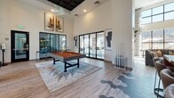 This single-floor amenity space is thoughtfully designed to offer a variety of functional zones, each with its own distinct character while maintaining a cohesive contemporary aesthetic throughout. Light wood-style flooring flows across much of the space, complemented by hexagonal tile accents that add visual interest at key transitions between areas.

The main lounge area features soaring ceilings and expansive floor-to-ceiling windows dressed in patterned drapes, flooding the space with natural light. A curved charcoal sectional sofa anchors the seating arrangement alongside woven accent chairs, a round coffee table, and a wall-mounted fireplace framed by a richly textured wood feature wall and built-in shelving. A bronze ring chandelier adds a refined finishing touch overhead.

An adjacent game room offers a full-size pool table beneath a dramatic double-height ceiling, with large glass sliding doors opening to an outdoor patio. Bold abstract artwork adorns the bright white walls throughout.

The kitchenette area makes a striking statement with a large-scale collage mural serving as a dramatic backdrop to dark cabinetry, stainless steel appliances, and a concrete-topped island with bar seating.

The co-working lounge features a long marble-topped conference table surrounded by vibrant yellow chairs, pendant lighting, individual desk stations along the windows, and a wall-mounted display screen—ideal for productive gatherings.