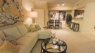 This single-floor space showcases an open-concept layout that seamlessly connects the living and dining areas, creating a cohesive and functional environment. The living area is anchored by a spacious sectional sofa in soft sage green, complemented by coordinating accent pillows in muted tones. A round glass-top coffee table with a dark metal frame serves as a stylish centerpiece, adorned with decorative candle holders and small accent pieces that add visual interest without overwhelming the space.

Warm, neutral wall tones throughout contribute to an inviting ambiance, enhanced by soft carpeting underfoot that adds comfort and continuity across the open floor plan. A floor lamp with a classic drum shade provides warm ambient lighting in the living area, while a track lighting fixture illuminates the dining space beyond.

The dining area features a dark-framed rectangular table paired with matching upholstered chairs, positioned beneath bold black-and-white artwork that serves as a striking focal point. A potted indoor tree adds a natural, organic element to the corner of the room. Crown molding details along the ceiling contribute an architectural refinement that elevates the overall design of the space.