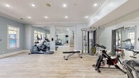 Located at 150 Carrington Park Dr in Gainesville, GA, this well-appointed fitness center offers a comprehensive workout environment spread across a single floor. The space is designed with a soft, calming palette of light blue walls, crisp white crown molding, and light wood-tone flooring that creates a bright, welcoming atmosphere throughout.

The facility is organized into multiple interconnected workout zones, allowing for a seamless flow between cardio and strength training areas. Large floor-to-ceiling mirrors line key sections of the space, visually expanding the room and providing exercisers with proper form feedback. Recessed lighting and ceiling fans maintain a comfortable and well-lit environment, while multiple windows along the perimeter invite natural light and views of the surrounding outdoor areas.

The cardio zone is equipped with elliptical trainers, treadmills, and stationary cycling bikes, offering a variety of aerobic workout options. The strength training area features a cable cross machine, a multi-functional weight station, and an adjustable workout bench, catering to a full range of resistance exercises. A dedicated room houses additional treadmill equipment alongside a wall-mounted television and a water cooler for added convenience.

Decorative touches such as a potted indoor tree add a refreshing, natural element to the space. The overall design balances functionality with an inviting aesthetic, making this fitness center a well-rounded facility for daily exercise routines.