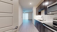 The E1B floor plan at Central Square at Frisco offers 480 square feet of efficiently designed living space on a single level. The galley-style kitchen is a standout feature, outfitted with dark espresso cabinetry, stainless steel appliances including an electric range, over-the-range microwave, dishwasher, and refrigerator, as well as light quartz countertops and a single-basin undermount sink. A white tile backsplash adds a clean, polished finish. Light wood-tone luxury vinyl plank flooring runs throughout the main living areas, complementing the neutral, warm color palette. Recessed lighting and a ceiling fan contribute to a bright, comfortable ambiance. The entryway provides a welcoming transition into the open living space. The bathroom features large-format tile flooring, a soaking tub and shower combination with full tile surround, and a toilet, offering a clean and functional layout. Directly accessible from the bathroom is a dedicated walk-in closet, equipped with built-in shelving, a hanging rod, and matching vinyl plank flooring, providing ample and organized storage. The apartment's layout maximizes every square foot, making it a practical and modern option for those seeking compact, well-appointed living at Central Square in Frisco.