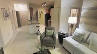 This single-floor residence showcases an open-concept layout that connects the living area and kitchen, creating a sense of spaciousness and ease of movement throughout. The living area is carpeted and comfortably accommodates seating arrangements, with a patterned accent chair, a rounded side table, and a sofa visible from the main vantage point. A tall floor lamp provides ambient lighting to complement the space. The kitchen is positioned toward the rear and features dark wood cabinetry, stainless steel appliances including a refrigerator and range, and dark stone-style countertops. A peninsula counter with bar stool seating serves as both a functional workspace and a casual dining spot, effectively dividing the kitchen from the living area. Track lighting on the ceiling illuminates the transition zone between the two spaces. Light-toned tile flooring in the kitchen contrasts with the carpeted living area, subtly defining each zone. A doorway visible near the kitchen hints at additional rooms beyond. The overall ambiance is modern and practical, with neutral tones and contemporary furnishings contributing to a clean, well-organized feel.