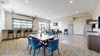 This single-floor community clubhouse is thoughtfully designed to serve as a versatile gathering space, blending modern aesthetics with functional layout. The grand lobby immediately impresses with soaring ceilings, exposed structural beams, and wide-plank wood-look flooring. A sleek linear fireplace is framed by a textured stone accent wall, while a striking black-framed barn door partition adds an industrial touch. Warm pendant lighting and cognac leather seating create an inviting atmosphere.

The main gathering area transitions seamlessly from carpet to hard flooring and accommodates multiple seating configurations, including dining tables with teal upholstered chairs, bar-height seating along a pass-through counter, and lounge clusters with dark leather sofas and armchairs. Wall-mounted flat-screen TVs are positioned throughout for entertainment. Large windows and glass doors allow generous natural light and offer views of surrounding green outdoor spaces.

The full kitchen is outfitted with stainless steel appliances, including a range, refrigerator, and built-in microwave, complemented by granite countertops, gray cabinetry, hardwood-style flooring, and a pass-through window to the lounge area—well-suited for hosting events and catered gatherings.