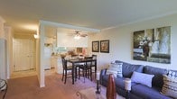 This single-story residence showcases an open-concept floor plan that seamlessly integrates the living, dining, and kitchen areas into one flowing, functional space. The living area is anchored by a comfortable sofa accented with decorative pillows, a glass-top coffee table, and framed wall art that adds a touch of personality to the neutral walls. Adjacent to the living space, the dining area features a dark wood table paired with matching high-back chairs, creating a defined yet connected zone for meals and gatherings. A ceiling fan with an integrated light fixture is centrally positioned, providing both air circulation and ambient lighting. The kitchen is partially open to the dining area and is defined by white cabinetry, a refrigerator, and a countertop that doubles as a casual workspace. Natural light filters in through the kitchen window, brightening the connected spaces. Soft carpeting runs throughout the main living and dining areas, contributing to a warm and comfortable atmosphere. Crown molding and a structural column add subtle architectural character to the overall design.