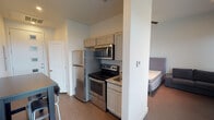 This well-designed studio unit offers a smart, open-concept floor plan that efficiently combines living, sleeping, and cooking areas into a single cohesive space. The kitchen is positioned along one wall and features stainless steel appliances, including a full-size refrigerator, an electric range, and an over-the-range microwave. Granite-look countertops, shaker-style cabinetry in a soft gray finish, and a compact sink complete the kitchen setup. A bar-height table with stools provides a dedicated dining area just steps from the kitchen.

The main living and sleeping area is bright and airy, benefiting from tall ceilings and multiple large windows that allow generous natural light to fill the room and offer open city views. The neutral color palette of white and light gray walls enhances the sense of space. Recessed lighting provides additional illumination throughout.

The sleeping area accommodates a full-size bed and features direct access to a private bathroom. The bathroom is clean and functional, equipped with a full-size soaking tub with an overhead shower, brushed nickel fixtures, and a glass shelf accent. Wood-look flooring flows consistently throughout the unit, lending warmth and continuity to the overall design. The entry door features decorative glass panel insets, adding a refined touch to the unit's modern aesthetic.