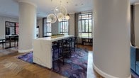 This elegantly designed single-floor space functions as a refined communal lounge, combining stylish aesthetics with practical functionality across an expansive, open layout. Structural columns positioned throughout the floor plan define distinct zones without fully separating them, creating an organic flow between areas dedicated to different uses.

The flooring throughout is a standout feature — rich herringbone-patterned hardwood that adds warmth and visual depth to the entire space. Vibrant, abstract-patterned area rugs in jewel tones of purple, blue, and mauve anchor the various seating clusters, adding contrast against the natural wood tones.

A central bar-height island with a striking marble surface serves as a social focal point, surrounded by sleek black bar stools and illuminated by dramatic starburst pendant chandeliers overhead. Nearby, low bookshelves line the walls beneath floor-to-ceiling steel-framed windows, which flood the interior with natural light and frame lush outdoor green views with sheer drape accents.

Gallery-style wall lighting installed in rows highlights the clean white walls, lending an art-forward atmosphere to one end of the space. Comfortable lounge seating — including upholstered armchairs and sofas with accent cushions — encourages relaxed conversation. High-top café tables near the entry provide additional informal gathering spots. The overall ambiance is polished, contemporary, and welcoming.