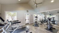 Located at 5000 Owens Dr in Pleasanton, CA, this fitness center is a well-appointed exercise space designed to support an active lifestyle. The room features striking vaulted ceilings that lend an open, airy feel despite the intimate footprint, with a ceiling fan providing steady air circulation during workouts.

The space is equipped with a solid selection of cardio machines, including treadmills, ellipticals, stationary bikes, and a rowing machine, all arranged to maximize usability. Light-toned hardwood-style flooring runs throughout, offering both durability and a clean aesthetic, while protective floor mats are positioned beneath the equipment.

Natural light pours in generously through multiple large windows framed in classic white trim, offering views of the surrounding greenery and creating an inviting atmosphere. A full-length mirror on one wall visually expands the space and allows users to monitor their form during exercise. A wall-mounted television provides entertainment for those on cardio machines.

Decorative touches, including an ornamental wall piece and a glass block accent panel, add character to the otherwise functional space. The neutral gray and white color palette keeps the environment calm and focused. Entry is accessible through both a glass-panel door and a secondary door, making the space easy to access from multiple points.