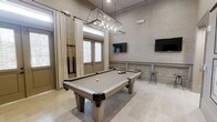This single-floor game room is thoughtfully designed with an upscale aesthetic that balances function and style. The centerpiece is a full-size billiard table finished in neutral tones, complemented by a striking geometric glass pendant light overhead that provides focused illumination while serving as a visual focal point. The room features textured, fabric-like wall coverings in soft greige tones, accented by classic wainscoting panels that lend a refined, traditional character. Two wall-mounted flat-screen televisions are positioned on opposite walls, ensuring excellent viewing angles from multiple vantage points throughout the space. Tall ceilings enhance the sense of openness, while frosted glass double doors and elevated transom windows allow diffused natural light to fill the room without compromising privacy. Long, floor-to-ceiling drapery panels frame the entry doors, adding a soft, tailored contrast to the otherwise structured design. Large-format neutral tile flooring ties the space together seamlessly. A cue rack and bar-height stools complete the room, making it a well-appointed destination for entertainment and leisure.