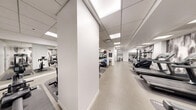 Located at 200 E Chestnut St in Chicago's prestigious Gold Coast neighborhood, this fitness center offers a generously sized, single-floor exercise space designed to support a comprehensive workout experience. The room is thoughtfully laid out to accommodate a wide variety of equipment, including multiple treadmills, elliptical trainers, a stair climber, and an assortment of strength and resistance machines, providing options for both cardiovascular and strength training.

The space features a clean, modern aesthetic defined by a neutral color palette of whites and grays, complemented by a striking large-scale abstract mural that serves as a dramatic focal point along one wall. Full-length mirrors on another wall visually expand the space and allow users to monitor their form during workouts. Overhead, a drop ceiling with recessed panel lighting provides bright, even illumination throughout.

Durable, speckled rubberized flooring covers the entire area, offering both safety and comfort underfoot. The open floor plan ensures ample room to move between equipment with ease. Subtle design details, such as sleek stainless steel waste receptacles, contribute to the polished, professional atmosphere. Fitness center guidelines are posted for user convenience, rounding out a well-organized and fully equipped facility.