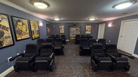 This dedicated screening room is thoughtfully designed to deliver a true theater experience in a comfortable, intimate setting. The room is finished with deep blue-gray walls accented by crisp white crown molding and baseboards, creating a refined yet cozy cinematic atmosphere. Patterned carpet in warm earth tones lines the floor throughout, adding texture and absorbing sound for an enhanced viewing experience.

Rows of black upholstered recliner chairs with built-in cup holders are arranged in a gentle arc facing a large flat-screen television mounted on the front wall, which is accompanied by a wood media console housing AV equipment. The seating capacity comfortably accommodates a sizeable group, making this space well-suited for communal movie screenings or private events.

A prominent wooden bookcase along the back wall houses an extensive collection of DVDs and media titles, offering a curated selection for viewers. The walls are adorned with a gallery of framed vintage classic movie posters — including iconic titles such as King Kong, Casablanca, The Wizard of Oz, and Gone with the Wind — lending the room a nostalgic, old-Hollywood charm.

Overhead, flush-mount circular ceiling fixtures provide warm ambient lighting, complemented by wall sconces that add a soft, theater-like glow. Multiple entry doors provide convenient access, and the overall layout ensures clear sightlines from every seat.