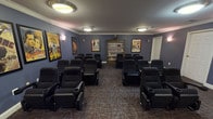 This dedicated screening room is thoughtfully designed to deliver a true theater experience in a comfortable, intimate setting. The room is finished with deep blue-gray walls accented by crisp white crown molding and baseboards, creating a refined yet cozy cinematic atmosphere. Patterned carpet in warm earth tones lines the floor throughout, adding texture and absorbing sound for an enhanced viewing experience.

Rows of black upholstered recliner chairs with built-in cup holders are arranged in a gentle arc facing a large flat-screen television mounted on the front wall, which is accompanied by a wood media console housing AV equipment. The seating capacity comfortably accommodates a sizeable group, making this space well-suited for communal movie screenings or private events.

A prominent wooden bookcase along the back wall houses an extensive collection of DVDs and media titles, offering a curated selection for viewers. The walls are adorned with a gallery of framed vintage classic movie posters — including iconic titles such as King Kong, Casablanca, The Wizard of Oz, and Gone with the Wind — lending the room a nostalgic, old-Hollywood charm.

Overhead, flush-mount circular ceiling fixtures provide warm ambient lighting, complemented by wall sconces that add a soft, theater-like glow. Multiple entry doors provide convenient access, and the overall layout ensures clear sightlines from every seat.