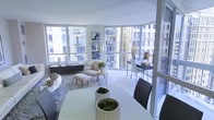 This single-floor space showcases a seamlessly open layout that integrates the living and dining areas into one cohesive, light-filled environment. The most defining architectural feature is the expansive floor-to-ceiling windows that wrap around the perimeter, flooding the interior with abundant natural light and offering unobstructed views of the surrounding urban cityscape. The generous glazing creates a strong visual connection between the indoors and the city beyond.

The living area is styled with a curved, cream-toned sofa paired with a matching ottoman and a geometric side table, all arranged atop a textured area rug. Framed artwork adorns one wall, adding a curated touch to the otherwise minimalist aesthetic. Adjacent to the living space, a sleek workspace or console table with modern chairs provides a functional secondary zone.

The dining area, positioned near the windows, features a clean white table accompanied by contemporary dark upholstered chairs, grounded by the same neutral flooring that runs throughout. Decorative elements, including moss ball centerpieces and sculptural vessels, contribute warmth and texture. The overall ambiance is refined, airy, and distinctly modern.