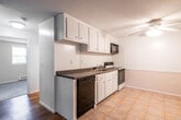 This single-floor residential unit offers a straightforward, functional layout with a variety of distinct living spaces. The open kitchen is well-equipped with white cabinetry extending to the ceiling for ample storage, dark countertops, a stainless steel sink, a white electric range, a black dishwasher, and an over-the-range microwave. A classic white tile backsplash adds a clean, timeless detail to the cooking area. The kitchen flows into an adjoining living area finished with light hardwood flooring, creating a bright and cohesive common space. A hallway connects the main living area to the rest of the unit, where warm hardwood flooring continues and a window draws in natural light. The bedroom features plush neutral carpet, a wall-mounted air conditioning unit, baseboard heating, and a window overlooking greenery outside. An additional carpeted room with two windows and baseboard heating provides flexible use as a second bedroom or office. The bathroom is finished with full white square tile surrounding the bathtub and shower combination, complemented by chrome fixtures. Throughout, textured ceilings and clean white walls create a neutral, move-in ready environment.