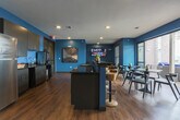 Located at 1700 Broad St in Chattanooga, Tennessee, this single-floor community space is defined by its bold teal and deep navy accent walls, warm hardwood flooring, and a cohesive modern design throughout. The lounge area provides a comfortable retreat, furnished with wood-framed black leather chairs arranged around a faceted gold coffee table, anchored by a large wall-mounted flat-screen television — ideal for relaxation or casual gatherings. Adjacent to the lounge, the fully equipped kitchen is outfitted with dark cabinetry, granite countertops, stainless steel appliances including a full-size refrigerator, a double sink, and upper storage cabinets. A breakfast bar with natural wood stools extends from the kitchen, offering an informal seating option. The dining area transitions seamlessly toward the outdoors through expansive floor-to-ceiling glass doors, flooding the space with natural light and opening onto a covered terrace. The terrace offers panoramic views of the Chattanooga skyline and surrounding landscape, with additional seating available for outdoor enjoyment. The overall ambiance is contemporary and energetic, with the vibrant color palette and thoughtful furnishings creating an inviting and functional environment throughout.
