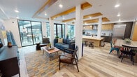Located at 2200 W Meeker St in Kent, Washington, this single-floor amenity space showcases a thoughtfully designed open-plan layout that seamlessly integrates a lounge, kitchen, and dining area. Distinctive warm wood beam accents contrast against crisp white ceilings, lending the interior a modern yet inviting character. Light-toned wood-style flooring runs throughout, complementing the airy, well-lit atmosphere created by large floor-to-ceiling windows and sliding glass doors that connect to an outdoor patio area.

The lounge features comfortable seating arrangements anchored by colorful area rugs, a wall-mounted flat-screen television, and vibrant original artwork that adds personality to the space. The fully equipped kitchen is a standout feature, offering stainless steel appliances including double wall ovens and a full-sized refrigerator, marble-style backsplash, a combination of dark and light wood cabinetry, and a generous island with butcher block countertops and bar seating beneath pendant lights. A dedicated dining area features a communal wood table surrounded by colorful mixed chairs, set against a striking blue reclaimed wood accent wall displaying coordinated framed artwork. Together, these elements create a dynamic, functional, and visually engaging communal environment.