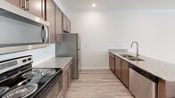This single-floor unit offers a well-organized layout with distinct living, sleeping, and utility areas finished in a clean, neutral palette throughout. The kitchen is a standout feature, equipped with stainless steel appliances including a refrigerator, range, microwave, and dishwasher. Rich dark brown cabinetry lines both walls of the galley-style kitchen, complemented by granite countertops and a white subway tile backsplash. A peninsula with an integrated sink and additional granite counter space extends into the adjoining area, providing a functional transition between the kitchen and living space. Light wood-look flooring runs throughout the main living areas, contributing to a bright, cohesive aesthetic.

A glass door with transom window in the living area allows natural light to flood the space, creating an airy atmosphere. Crown molding adds a refined detail to the main living areas. The bedroom features soft wall-to-wall carpeting and a window with blinds that brings in natural light, while a dedicated walk-in closet with wire shelving provides generous storage. The in-unit laundry room includes a full-size washer and dryer alongside additional wire shelving, offering practical everyday convenience. Recessed lighting and consistent neutral wall tones unify the overall interior.