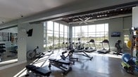This single-floor fitness space is defined by its striking wall of floor-to-ceiling windows, which floods the interior with natural light and offers expansive views of the surrounding outdoor landscape. The open layout provides ample room for a comprehensive selection of exercise equipment, including treadmills, elliptical machines, rowing machines, and adjustable weight benches, all arranged to maximize usability and circulation throughout the space.

The ceiling features exposed industrial ductwork alongside a large ceiling fan, contributing to a modern, utilitarian aesthetic while supporting airflow and ventilation across the workout area. Polished concrete flooring runs throughout, offering a durable and easy-to-maintain surface well-suited to a high-activity environment. A wall-mounted television is positioned for visibility from multiple workout stations.

The abundance of natural light, combined with the exterior views of trees and open grounds, creates an energizing and visually open atmosphere. The overall design balances functional fitness infrastructure with a clean, contemporary aesthetic, making it a well-equipped and comfortable environment for a full range of exercise activities.