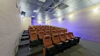 This single-floor private cinema screening room is designed to deliver an authentic movie theater experience. The auditorium is anchored by a large projection screen at the front, dramatically framed by sweeping black stage curtains adorned with elegant gold trim detailing — a classic theatrical touch that sets the tone for the space. A ceiling-mounted projector is positioned centrally overhead, ensuring optimal image quality across the full width of the screen.

The seating area is arranged in gently tiered rows of cushioned, high-back theater chairs upholstered in rich rust-red fabric, each equipped with armrests and cup holders for comfort. A low dividing partition with LED accent lighting runs along one side of the auditorium, adding a modern visual element and helping to define the space. Side-aisle steps provide easy access between seating rows.

A surround sound speaker system is distributed throughout the room, with speakers mounted along the side walls and flanking the screen, supporting an immersive audio experience. The drop-panel ceiling features recessed and flush-panel lighting, while wall-mounted sconces provide warm ambient illumination. The dark carpeted flooring and muted wall tones contribute to the room's intimate, focused atmosphere, minimizing distractions and enhancing the overall viewing environment.