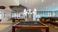 Located at 4555 McCrimmon Pkwy in Morrisville, NC, this single-floor clubhouse is a thoughtfully designed amenity space that blends social, recreational, and professional functionality. The open-concept main area serves as the heart of the space, featuring a full-size pool table beneath striking globe pendant lights with brass accents, complemented by herringbone-patterned carpet and light hardwood flooring. A stylish lounge area offers plush sectional seating and large black-framed windows that flood the room with natural light and views of the surrounding community. An adjacent kitchen bar is outfitted with white cabinetry, a large island, stainless steel appliances, hexagonal tile backsplash, and multiple mounted televisions. The business center provides dedicated workstations along a built-in desk, vibrant orange accent chairs, and glass-partitioned walls for a semi-open feel. A private conference room features a live-edge wood table, leather executive chairs, a wall-mounted display, and bold mustard-yellow walls adorned with decorative propeller artwork. Individual work pods with industrial-style mural backdrops offer focused workspace. The covered outdoor terrace, with a slatted wood ceiling, dining table, and lounge seating, overlooks a resort-style pool courtyard below.