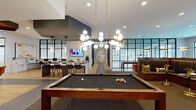 Located at 4555 McCrimmon Pkwy in Morrisville, NC, this single-floor clubhouse is a thoughtfully designed amenity space that blends social, recreational, and professional functionality. The open-concept main area serves as the heart of the space, featuring a full-size pool table beneath striking globe pendant lights with brass accents, complemented by herringbone-patterned carpet and light hardwood flooring. A stylish lounge area offers plush sectional seating and large black-framed windows that flood the room with natural light and views of the surrounding community. An adjacent kitchen bar is outfitted with white cabinetry, a large island, stainless steel appliances, hexagonal tile backsplash, and multiple mounted televisions. The business center provides dedicated workstations along a built-in desk, vibrant orange accent chairs, and glass-partitioned walls for a semi-open feel. A private conference room features a live-edge wood table, leather executive chairs, a wall-mounted display, and bold mustard-yellow walls adorned with decorative propeller artwork. Individual work pods with industrial-style mural backdrops offer focused workspace. The covered outdoor terrace, with a slatted wood ceiling, dining table, and lounge seating, overlooks a resort-style pool courtyard below.