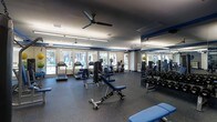 This single-floor fitness center offers a comprehensive workout environment designed to accommodate a range of exercise needs. The space is finished with durable rubber flooring in a speckled dark grey tone, providing both safety and functionality throughout. A dark ceiling with ceiling fans ensures air circulation, while recessed and strip lighting keeps the space well-lit.

Large windows along one wall flood the interior with natural light and offer views of the surrounding outdoor area, creating an open and inviting atmosphere. Mirrored walls on the opposite side visually expand the space and allow users to monitor their form during workouts.

The cardio zone features multiple treadmills and an elliptical trainer positioned near the windows, taking advantage of the natural light and views. The strength training area includes a cable machine, an adjustable incline bench, and a well-stocked dumbbell rack with weights neatly arranged in ascending order. Blue accents on the equipment and architectural trim tie the design together cohesively. The layout is spacious and thoughtfully organized, allowing for comfortable movement between stations.