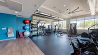 This single-floor fitness center is thoughtfully designed to accommodate a comprehensive workout experience. The space is divided into distinct functional zones, each purpose-built for different training disciplines. A dedicated Training Zone lines one wall, accented by a bold teal accent wall and stocked with a full rack of dumbbells, medicine balls, resistance bands, stability balls, and a multi-station cable machine. Floor-to-ceiling mirrors enhance the sense of space and allow users to monitor their form during exercises.

The cardio section is lined with large, floor-to-ceiling windows that bathe the room in natural light and offer open outdoor views, creating an airy and energizing atmosphere. This area houses treadmills, elliptical machines, stationary bikes, and cycling equipment, all arranged to take advantage of the scenic outlook. Exposed industrial-style ductwork runs along the ceiling alongside large ceiling fans, contributing to both ventilation and a modern aesthetic.

Durable rubber flooring covers the workout areas, transitioning to hardwood-style flooring near the entry and accessory zones. A hydration station with a bottle-fill water fountain is conveniently located within the space, accompanied by yoga mats and foam rollers stored nearby. Recessed lighting complements the abundant natural light, ensuring the space remains bright and welcoming throughout the day.