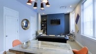 This single-floor space showcases a thoughtfully curated interior design that balances bold aesthetic choices with functional living. The main living area immediately draws the eye with a striking navy blue accent wall, which serves as a dramatic backdrop for a wall-mounted flat-screen television. Light blue walls throughout the remainder of the space provide a complementary contrast, lending the interior a cohesive and calming palette.

The living area is anchored by a sleek black leather sofa and a glass-topped coffee table that reflects the warm tones of the light wood flooring below. A statement multi-arm chandelier with dark cylindrical shades hangs overhead, adding an artistic focal point that enhances the contemporary character of the room. Recessed lighting further illuminates the space with a clean, modern touch.

A built-in bookshelf filled with books and decorative objects adds depth and personality to the room, while a round gold-framed mirror on the wall introduces an elegant, classic accent. A dedicated workspace or reading nook near the window features a chair and desk arrangement, taking advantage of natural light. The overall ambiance is polished and intentional, combining modern fixtures with rich textures and a carefully considered color story throughout.