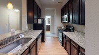 Located at 15705 Colony Oaks Dr in Charlotte, NC, this single-story residence offers a well-organized layout with a variety of functional spaces. The galley-style kitchen is a standout feature, showcasing dark espresso cabinetry, granite countertops with a tile backsplash, and black stainless steel appliances including a range, microwave, and refrigerator. Light hardwood-style flooring runs throughout the kitchen, complementing the rich cabinetry tones.

The living areas feature neutral-toned carpet and crown molding in select rooms, contributing to a polished, cohesive aesthetic. Multiple bedrooms are present, each carpeted and equipped with ceiling light fixtures and windows fitted with blinds that allow natural light to filter in while maintaining privacy. The rooms offer views of surrounding greenery, enhancing the sense of connection to the outdoors.

The bathrooms are finished with white tile tub-and-shower combinations, chrome fixtures, and dark vanity cabinetry topped with light countertops. Hardwood-style flooring in the bathrooms provides a clean, practical surface. A private exterior door in one room adds convenient access. Overall, the space presents a neutral, move-in-ready environment with consistent finishes throughout.